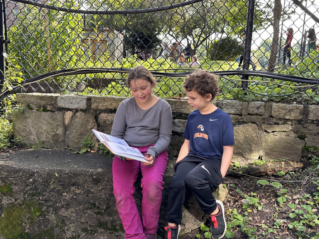 Two Fieldston Lower students read together while sitting on a rock in the outdoor garden.