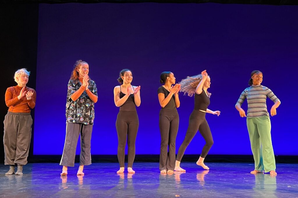Performers stand in a line on stage, some are clapping, others smiling.