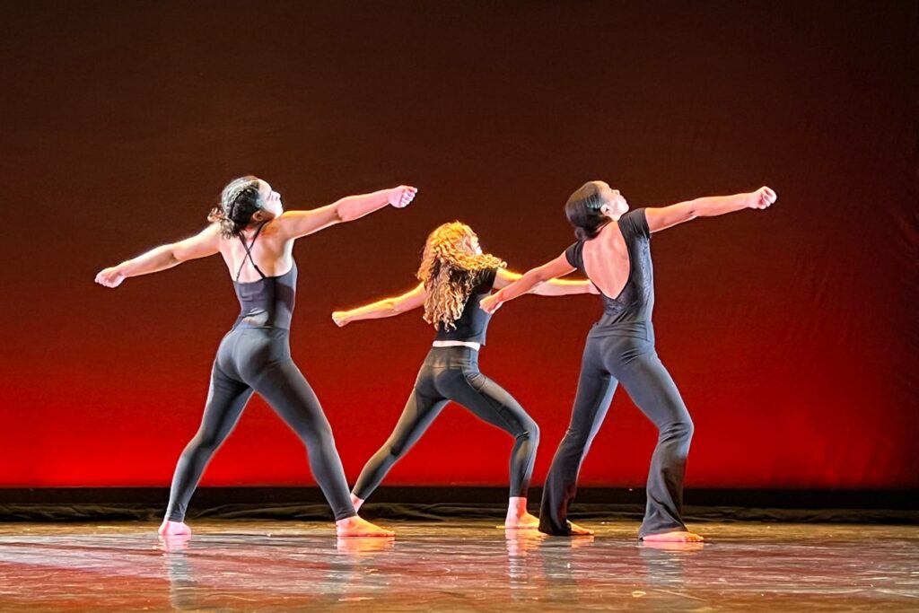 Three Fieldston Upper Dancers perform on stage, wearing all back and stretching their arms and legs out to the side.