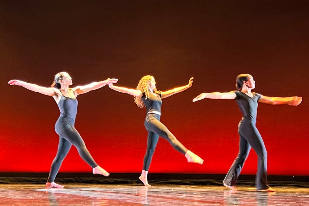 Three Fieldston Upper dancers perform on stage, wearing all black; the stage lighting is a dark red tone.