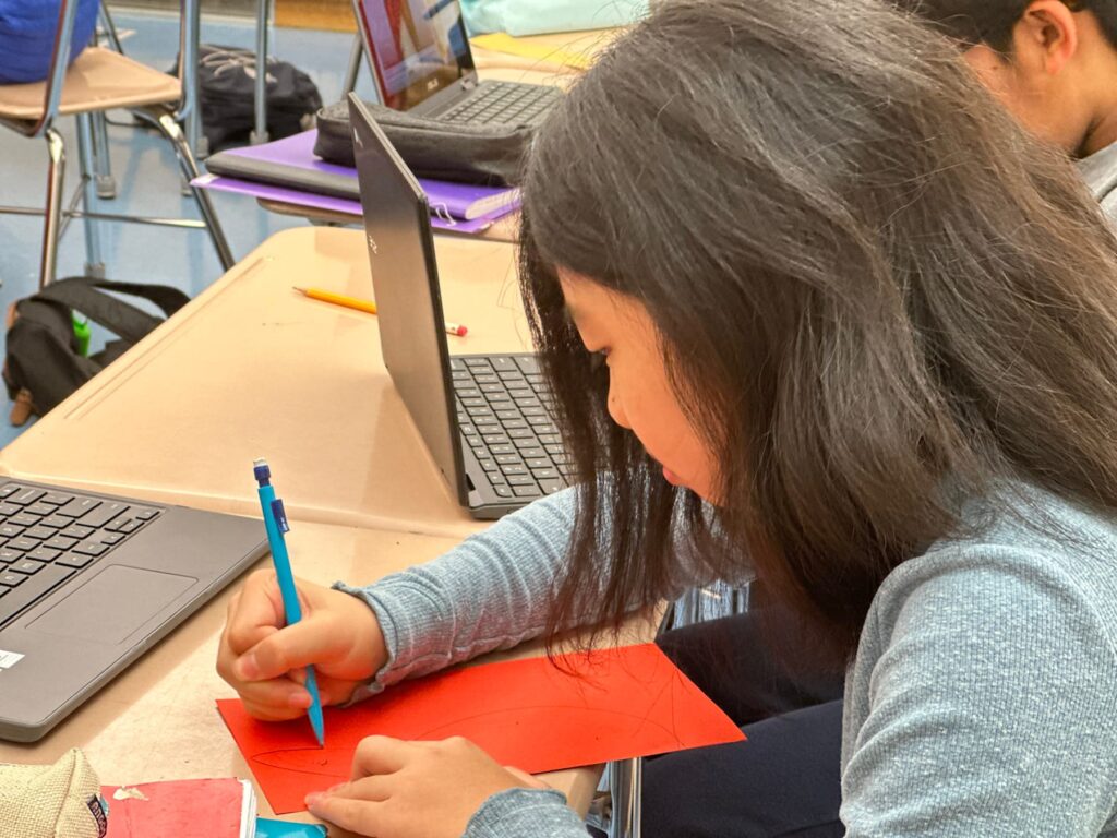 Student uses pencil to trace an outline on red paper.