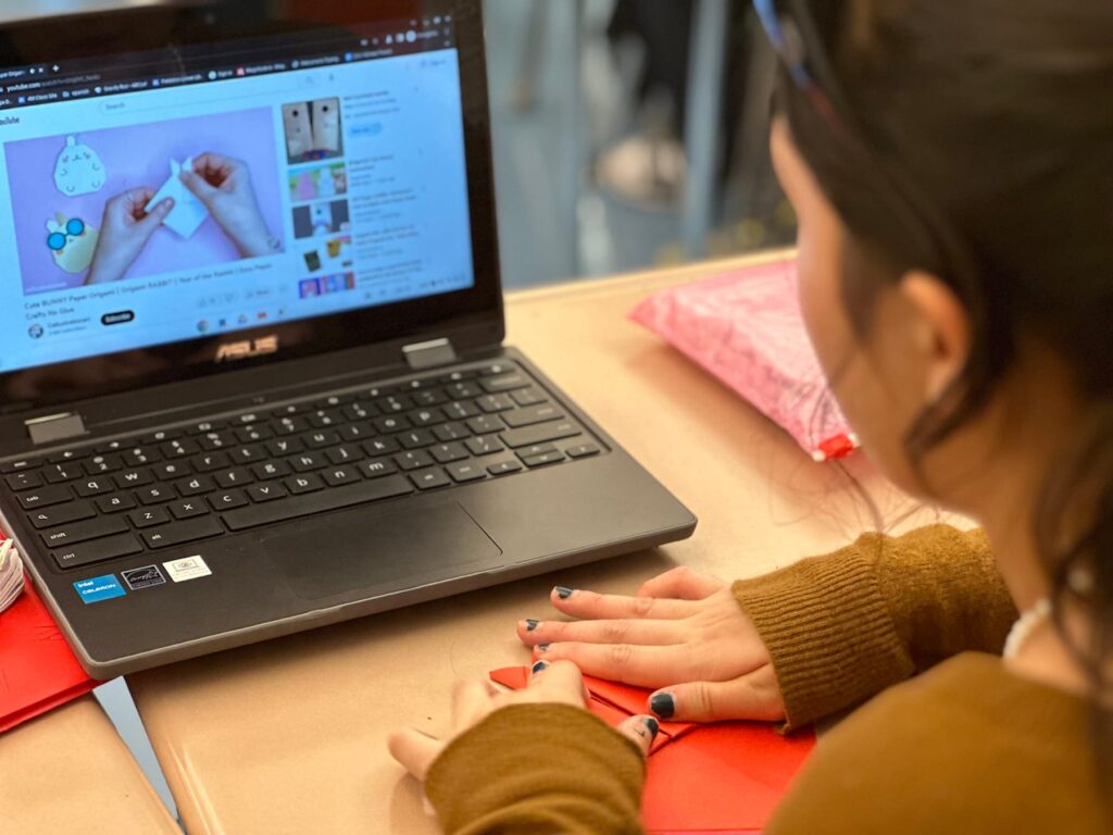 Student looks at computer tutorial while folding a rabbit origami.